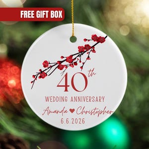 May include: A round white ceramic ornament featuring a red cherry blossom branch illustration. The text reads 40th Wedding Anniversary, Amanda and Christopher, and the date 6.6.2026 in red lettering. A gold string is attached to the top hole.