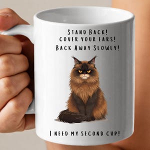 May include: White ceramic coffee mug with a black and brown cat illustration. The mug has the text "STAND BACK! COVER YOUR EARS! BACK AWAY SLOWLY! I NEED MY SECOND CUP!" printed on it.