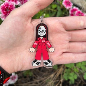 May include: A clear acrylic keychain featuring a cartoon character with long black hair and white face paint, wearing a red jumpsuit with a white number one on the chest, a barcode graphic, and a white logo on the sleeve, paired with white sneakers.