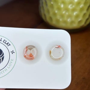 May include: Two round stud earrings with a clear resin finish, showcasing orange and white flecks. The earrings are displayed on a white card, with a blurred green vase in the background. The earrings are a jewelry item.
