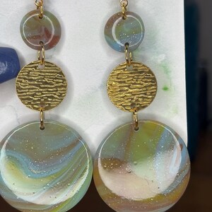 May include: A pair of dangle earrings with a gold-tone finish. Each earring features three circular elements: a small top circle, a textured gold disc, and a large bottom circle with a marbled effect in shades of green, blue, and brown.