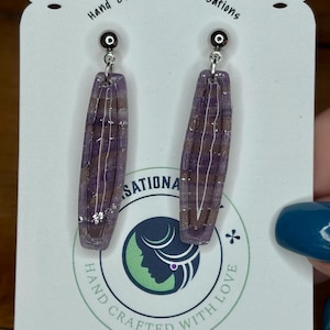 Pode incluir: Um par de brincos de argila artesanais com formato retangular. Os brincos são roxos translúcidos com detalhes em marrom e branco. Pendurados em pinos de metal prateado. O cartão branco tem o texto "Simsational Clay".
