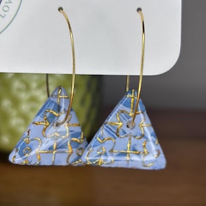 May include: A pair of earrings featuring light blue triangle pendants with gold floral patterns, suspended from gold-colored hoop wires. The earrings are displayed on a white card.