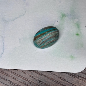 May include: An oval-shaped, handcrafted stone with a marbled design. The stone features bands of teal, brown, and gold, creating a unique pattern. The polished surface reflects light, highlighting the color variations. This decorative stone is ideal for jewelry making or as a decorative accent.