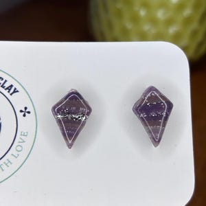 Könnte beinhalten: Ein Paar rautenförmige Ohrstecker. Die Ohrringe sind lila mit silbernen Glitzerakzenten. Sie werden auf einer weißen Karte präsentiert. Der Hintergrund ist verschwommen, mit einem grünen und braunen Farbschema.