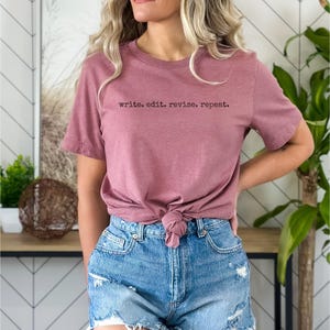 Könnte beinhalten: Ein staubrosa T-Shirt mit dem Text "write. edit. revise. repeat." in Schwarz. Das Shirt ist an der Taille geknotet und wird mit Jeansshorts kombiniert. Der Hintergrund zeigt eine dekorative Wand und Grünpflanzen.