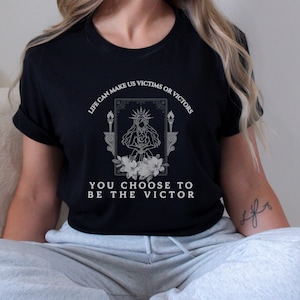 May include: Black t-shirt with white text and graphic. The graphic features a crowned figure with a crescent moon, surrounded by flowers and candles. Text reads "LIFE CAN MAKE US VICTIMS OR VICTORS YOU CHOOSE TO BE THE VICTOR."