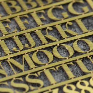 May include: Close-up of a sheet of gold-coloured metal letters and numbers. The letters are in a sans-serif font and are arranged in rows. The numbers include 8, 9, 0, 5, and 4. The background is a textured grey surface.
