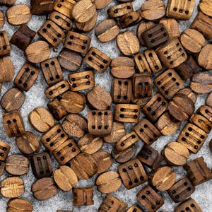 May include: A collection of small, wooden beads in various shapes and shades of brown. Some beads are oval, while others are rectangular with carved details. The beads are scattered on a light grey surface.