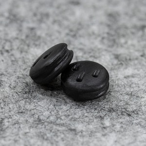 May include: Two black buttons with a matte finish sit on a grey, textured surface. One button is oval-shaped with a single hole, the other is round with three small holes. These buttons are likely for clothing or crafts.