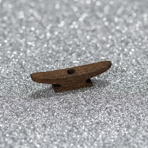 Puede incluir: Un pequeño objeto marrón en forma de yunque con un agujero en el centro. El objeto está hecho de un material texturizado y se encuentra sobre una superficie plateada brillante. El yunque tiene una parte superior plana y una base con dos pequeños soportes.