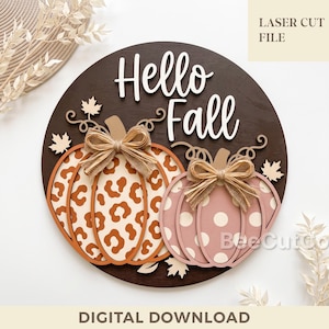 Puede incluir: Un letrero circular de madera marrón oscuro con el texto blanco Hello Fall. Dos calabazas superpuestas están fijadas en la parte frontal, una con estampado de leopardo marrón y la otra con lunares rosas, cada una decorada con un lazo de arpillera.