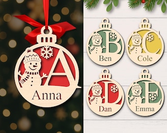 May include: Wooden Christmas ornaments with snowman and initial designs. One ornament has the name "Anna" and a red background. Other ornaments feature the names "Ben", "Cole", "Dan", and "Emma". Each ornament has a snowman design.