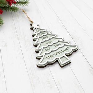 May include: A wooden Christmas tree ornament with a light gray and brown layered design. The ornament is shaped like a Christmas tree and features the names "Liam", "Olivia", "Noah", "Dad", "Mom", and "THE WILLIAMS". The year "2025" is also included.