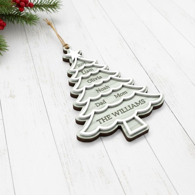 May include: A wooden Christmas tree ornament with a light gray and brown layered design. The ornament is shaped like a Christmas tree and features the names "Liam", "Olivia", "Noah", "Dad", "Mom", and "THE WILLIAMS". The year "2025" is also included.