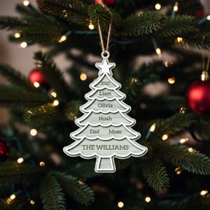May include: A Christmas tree-shaped ornament with a star on top, featuring the year 2025, family names, and "THE WILLIAMS" at the bottom. The ornament is white with a light gray interior and hangs from a brown string. The background is a decorated Christmas tree.
