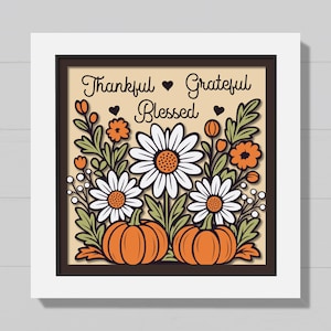 May include: Framed artwork with the words "Thankful, Grateful, Blessed" above a floral and pumpkin design. The artwork features white daisies, orange flowers, green leaves, and two orange pumpkins. The frame is dark brown.