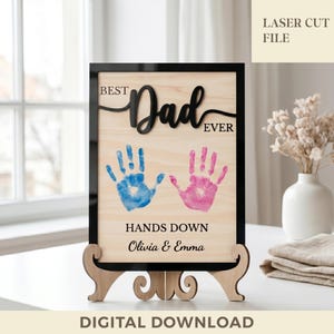May include: Wooden sign with a black frame and stand, featuring the words "Best Dad Ever" in black script. Below are blue and pink handprints, with the text "Hands Down Olivia & Emma". The words "Laser Cut File" and "Digital Download" are also visible.