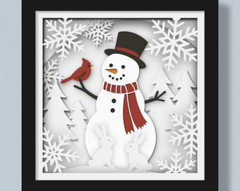 May include: A framed paper art piece featuring a snowman with a black top hat and a red scarf, a red cardinal, and two white rabbits. The background includes white snowflakes and trees, all within a black frame.