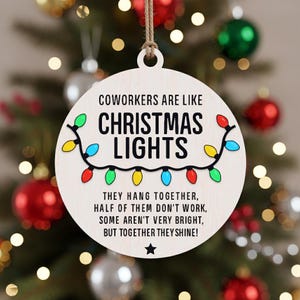 May include: A round, wooden Christmas ornament with the text "COWORKERS ARE LIKE CHRISTMAS LIGHTS." The ornament features a string of colorful lights and the text "THEY HANG TOGETHER, HALF OF THEM DON'T WORK, SOME AREN'T VERY BRIGHT, BUT TOGETHER THEY SHINE!"