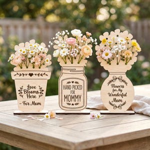 Puede incluir: Tres portaflores de madera con arreglos florales. Los portaflores tienen forma de maceta, tarro y jarrón. Cada portaflores tiene el texto: "Love Blooms Here - For Mom", "Hand Picked For Mommy" y "Flowers for My Wonderful Mom".