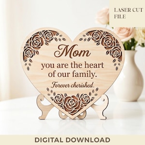Puede incluir: Un letrero de madera en forma de corazón con diseños florales cortados con láser y las palabras "Mamá, eres el corazón de nuestra familia. Siempre apreciada." El letrero está sostenido por un soporte de madera. Las palabras "LASER CUT FILE" y "DESCARGA DIGITAL" también son visibles.