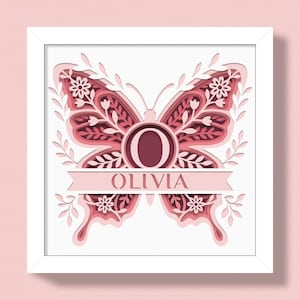 May include: A framed, layered paper art piece featuring a butterfly design in shades of pink and red. The butterfly has floral and leaf details, with the name "OLIVIA" displayed on a banner. The letter "O" is in the center.