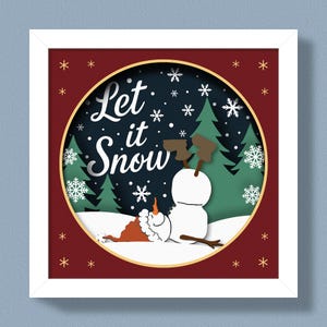 Puede incluir: Una obra de arte enmarcada con temática invernal. La obra presenta un muñeco de nieve con brazos de ramitas y una nariz de zanahoria, tumbado en la nieve. Las palabras "Let it Snow" están escritas en blanco. El fondo incluye copos de nieve y pinos.