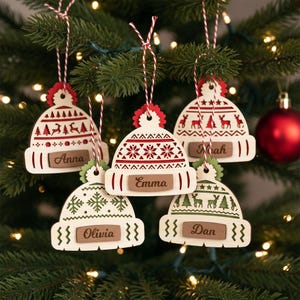 Puede incluir: Adornos navideños de madera con forma de gorros, cada uno con una etiqueta con nombre. Los gorros blancos presentan patrones rojos y verdes de copos de nieve, árboles y renos. Cuelgan de un árbol de Navidad con hilo rojo y blanco.