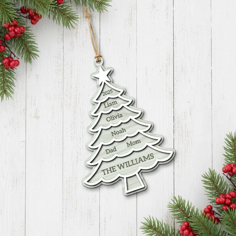 May include: A light gray wooden Christmas tree ornament with a star on top and the year 2025. The ornament is engraved with names and the family name "THE WILLIAMS". It hangs from a natural twine string.