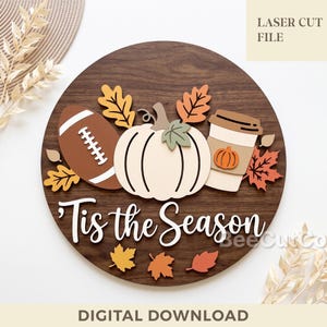 May include: A circular wooden sign featuring a dark brown wood grain background with layered cutouts of a brown football, a cream-colored pumpkin, and a coffee cup. The text 'Tis the Season' is in white script. Includes orange and yellow leaf accents.