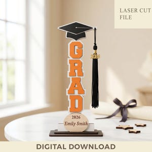Puede incluir: Decoración de graduación de madera con la palabra "GRAD" en letras naranjas. Cuenta con un birrete negro, una borla negra con un dije dorado de 2026 y una base de madera con un nombre y un año. También se ven las palabras "LASER CUT FILE" y "DIGITAL DOWNLOAD".