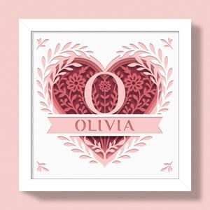 Op de afbeelding: Een ingelijst papieren kunstwerk met een hartontwerp, bloemen en bladeren. De naam "OLIVIA" staat op een banner. De letter "O" staat in het midden van het hart, met een bloemenontwerp. Het kunstwerk is in roze tinten.