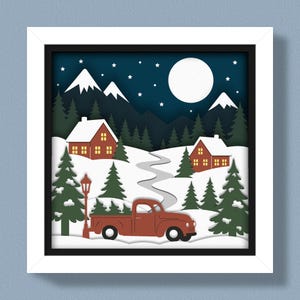 May include: A framed paper art piece depicting a winter scene. A red pickup truck carries a Christmas tree on a snowy road, passing by snow-covered houses and evergreen trees. The night sky features a full moon and stars.