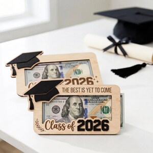 May include: Wooden graduation gift frames with a black mortarboard design, holding a $100 bill. The frames are engraved with "Class of 2026" and "The Best Is Yet To Come". A graduation cap and diploma are in the background.