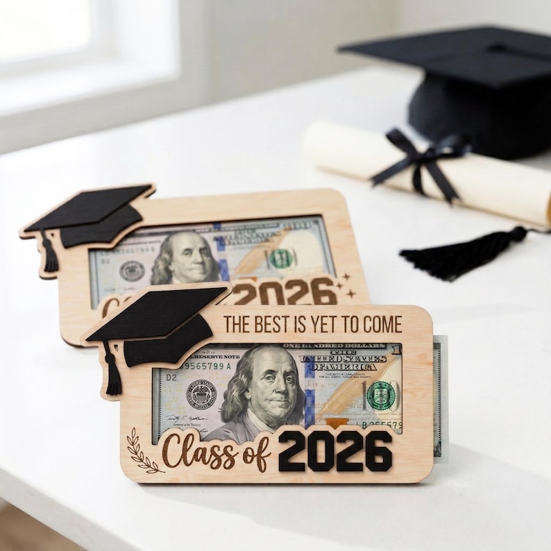 May include: Wooden graduation gift frames with a black mortarboard design, holding a $100 bill. The frames are engraved with "Class of 2026" and "The Best Is Yet To Come". A graduation cap and diploma are in the background.