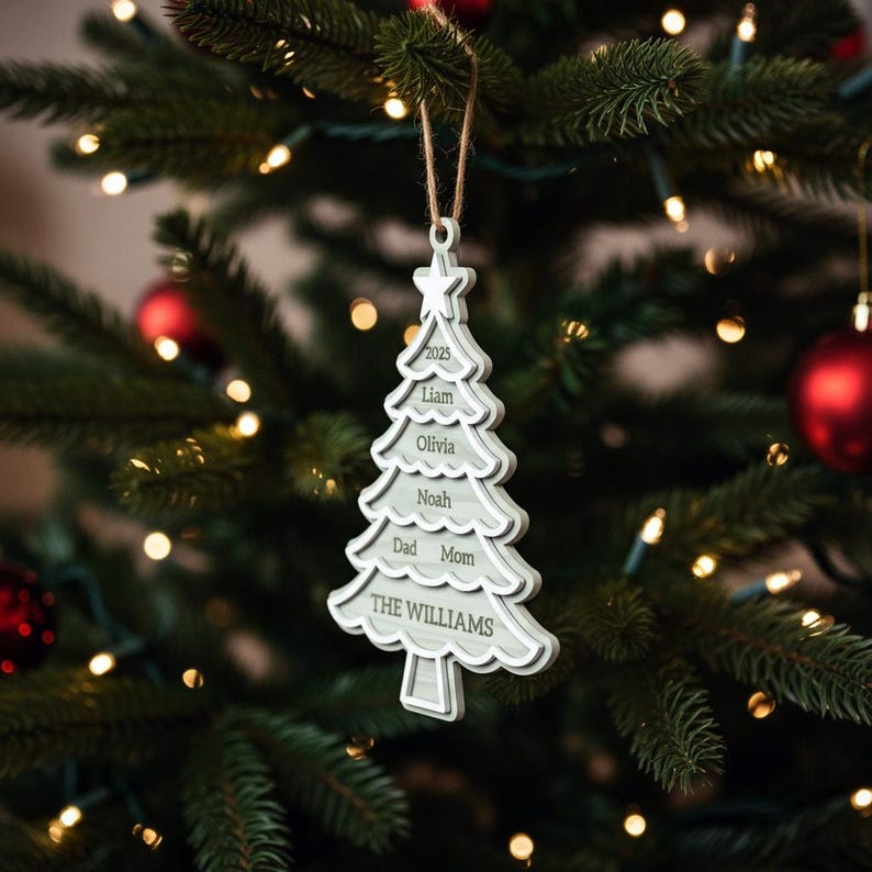 May include: A white and gray Christmas tree ornament with the names Liam, Olivia, Noah, Dad, and Mom, and the family name "THE WILLIAMS" printed on it. The ornament is hanging on a Christmas tree.