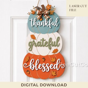 May include: A vertical wall hanging featuring three stacked wooden pumpkins in teal, white, and orange. The pumpkins are decorated with small wooden leaves, acorns, and a plaid bow. The words thankful, grateful, and blessed are written in script.