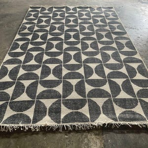 Könnte beinhalten: Ein rechteckiger Teppich mit einem sich wiederholenden geometrischen Muster aus dunkel anthrazitfarbenen Halbkreisen auf cremefarbenem Grund. Der Teppich hat eine gewebte Textur und ist an den beiden kurzen Seiten mit kurzen, hellen Fransen versehen.