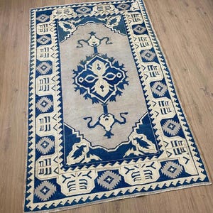 Könnte beinhalten: Rechteckiger Teppich mit einem zentralen Design in Blau und Grau, umgeben von einem Rand aus geometrischen Mustern in Blau und Creme. Das zentrale Feld des Teppichs ist hellgrau. Der Teppich liegt auf einem Holzboden.