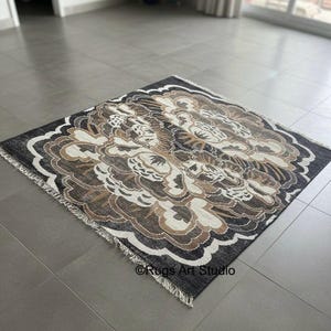 Peut inclure: Un tapis carré avec un motif floral dans les tons de noir, marron et crème. Le tapis a une bordure à franges et un motif détaillé. Le texte "Rugs Art Studio" est visible dans le coin inférieur.