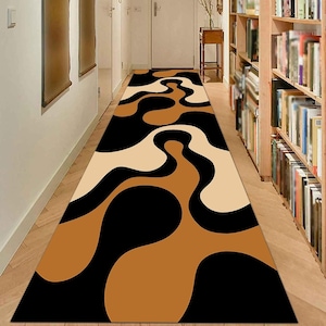 Könnte beinhalten: Ein rechteckiger Teppichläufer mit einem abstrakten Design in Schwarz, Beige und Braun. Das Muster weist fließende, organische Formen auf, die einen visuell interessanten Kontrast erzeugen. Der Teppich liegt in einem Flur mit Holzboden und Bücherregalen.