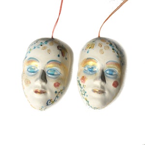 May include: Two white ceramic masks with painted details. Each mask has blue eyes, gold accents, and floral designs in shades of pink, blue, and green.