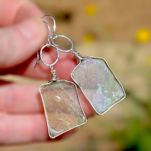 May include: A pair of dangle earrings featuring rectangular pieces of iridescent, semi-translucent glass wrapped in silver-toned wire. Each earring hangs from a silver-toned circular link and a curved ear wire, showcasing a shimmering, multi-colored surface.