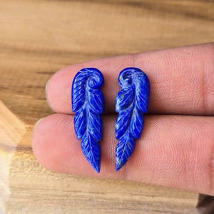 Pode incluir: Um par de esculturas de pedra preciosa lápis-lazúli azul profundo em forma de penas alongadas. Cada peça apresenta detalhes intrincados de veios esculpidos na superfície, destacando a textura natural e a cor azul vibrante e saturada da pedra.