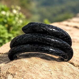 Könnte beinhalten: Ein glänzend schwarzes Armband in Form einer gewundenen Schlange mit einem detaillierten, texturierten Schuppenmuster, das die gesamte Oberfläche bedeckt. Das Design zeigt einen glatten, abgerundeten Kopf auf den Windungen des Schlangenkörpers.