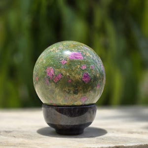 May include: A polished spherical stone featuring a mottled green base with embedded vibrant pink mineral inclusions and small gold-colored flecks, resting securely on a smooth, dark gray, bowl-shaped stone stand.