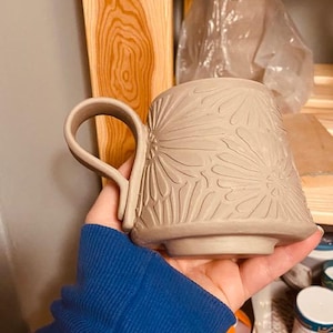 May include: A light beige ceramic mug featuring a carved floral pattern across its exterior surface. The mug has a wide, curved handle attached to the side and a raised circular base. The clay surface is matte and shows intricate petal details.