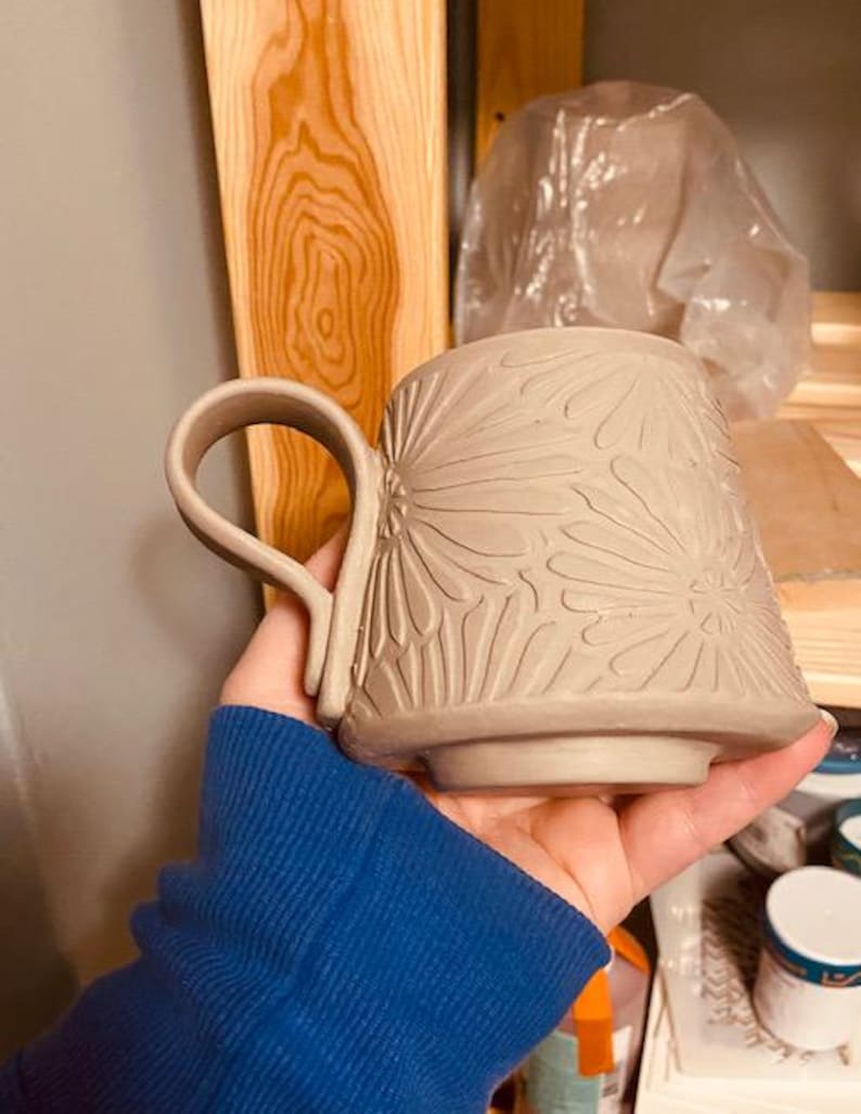 May include: A light beige ceramic mug featuring a carved floral pattern across its exterior surface. The mug has a wide, curved handle attached to the side and a raised circular base. The clay surface is matte and shows intricate petal details.
