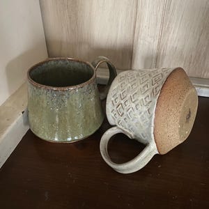 May include: Two ceramic mugs. One mug is upright with a mottled olive green glaze and a smooth finish. The second mug is lying on its side, featuring a cream-colored exterior with a raised geometric diamond pattern and a textured brown base.